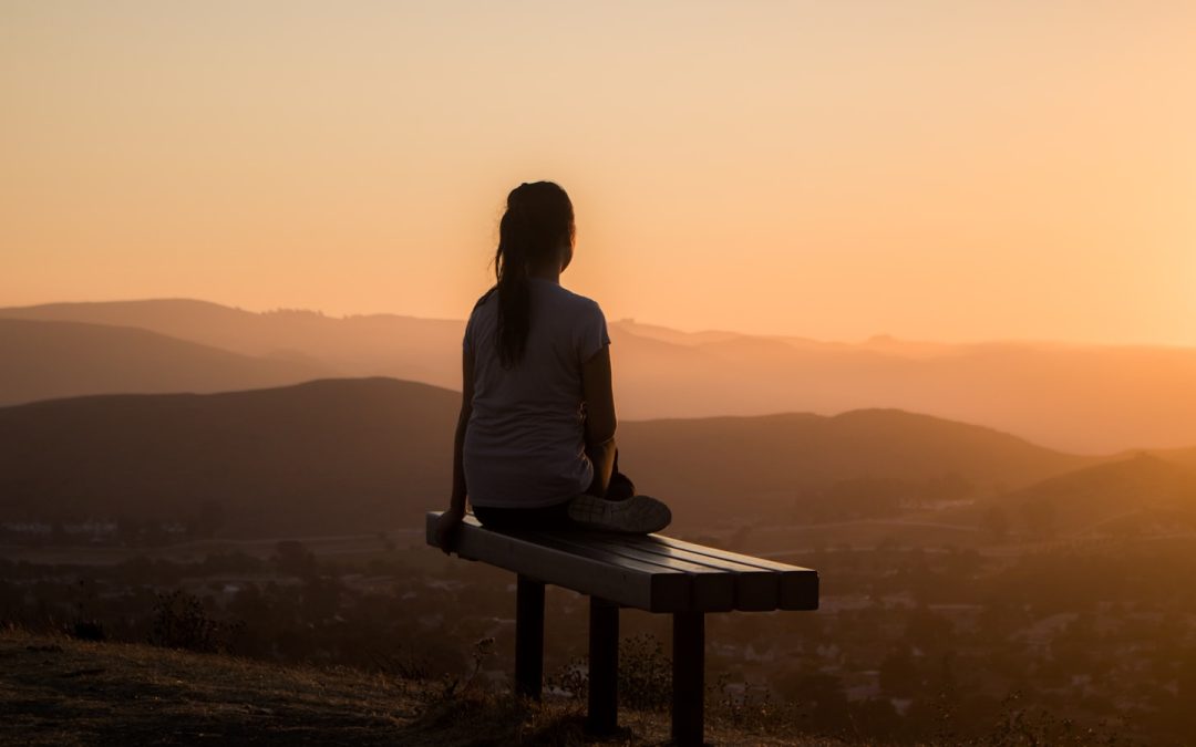 Woman sitting alone on a bench at sunset, looking out over hills, representing rest and recovery from burnout