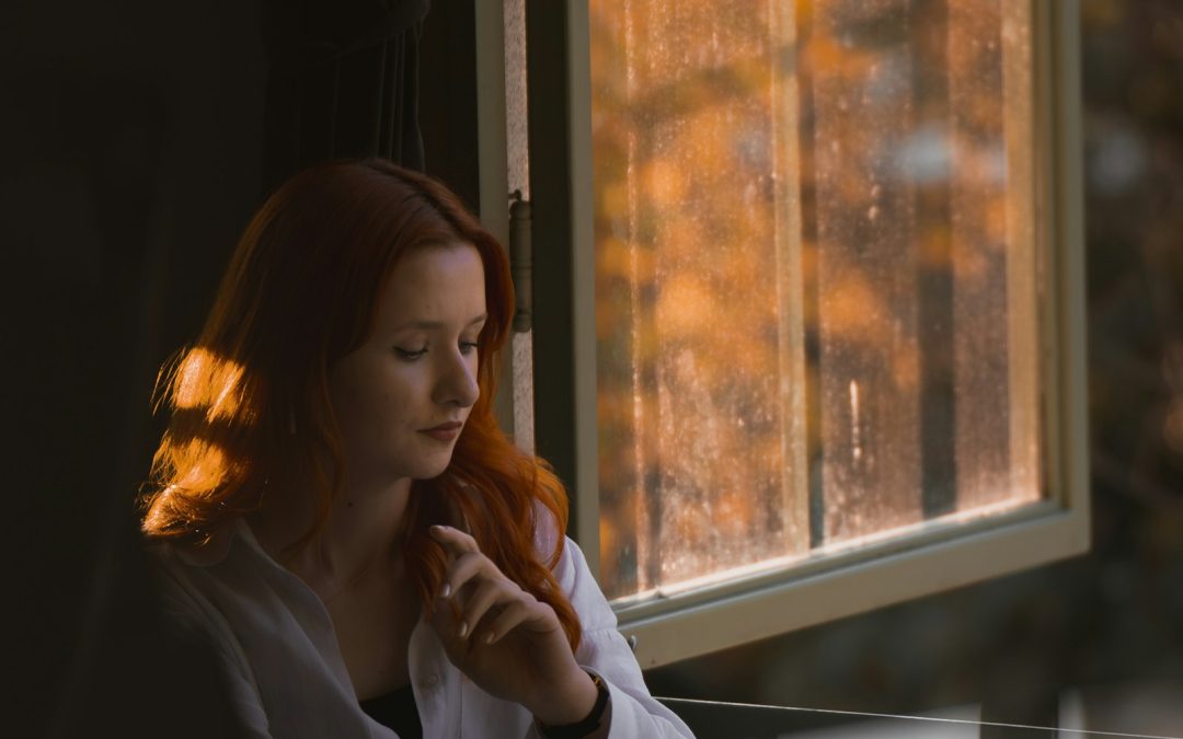 Woman sitting quietly by a window in warm autumn light, reflecting during the end-of-summer transition into September.