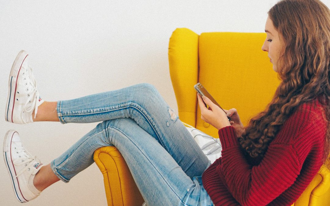 Person relaxing on a chair while looking thoughtfully at a smartphone, representing the choice between AI chatbot support and in-person therapy
